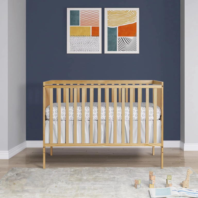 Natural-toned wooden crib with vertical slats and straight rails pictured in front of a dark blue wall in a furnished room